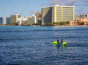 A few people await a wave while surfing, with two volunteers in highlighter colored rash guards. The view of Honolulu buildings shows in the background. 